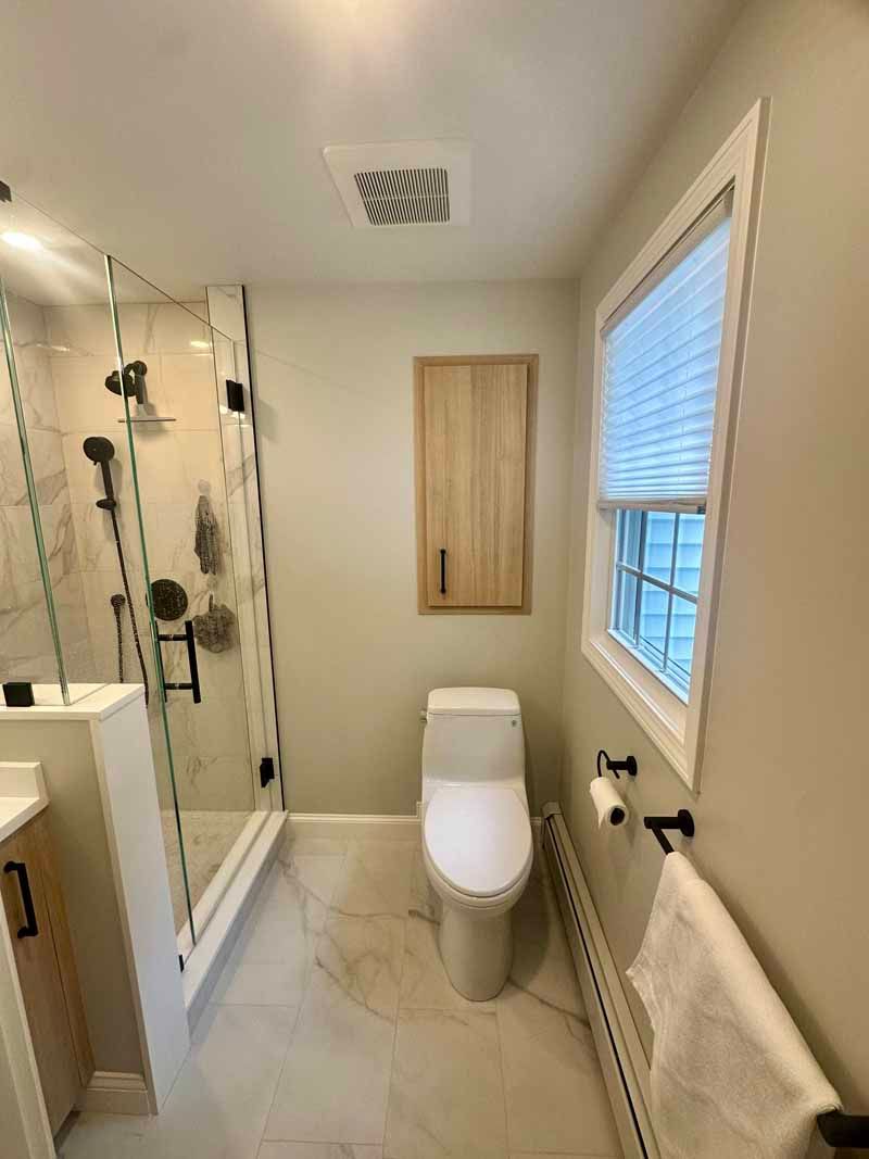 A remodeled bathroom with a glass shower, toilet, and a window with blinds.
