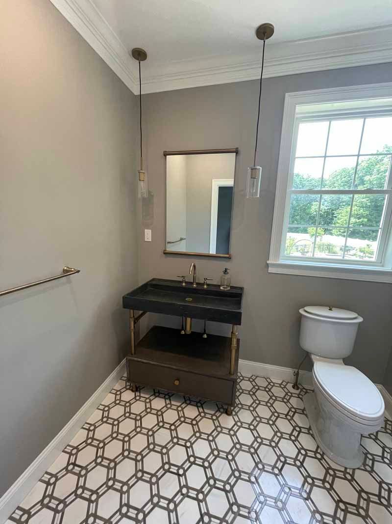 Bathroom with patterned floor, dark vanity, mirror, toilet, and two hanging lights. Gray walls, window.