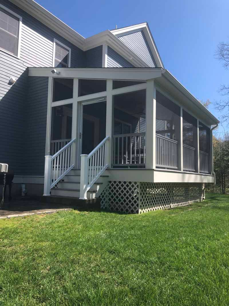 Screened-in white porch attached to a blue house, featuring steps, railing, and lattice skirting, set on green grass.