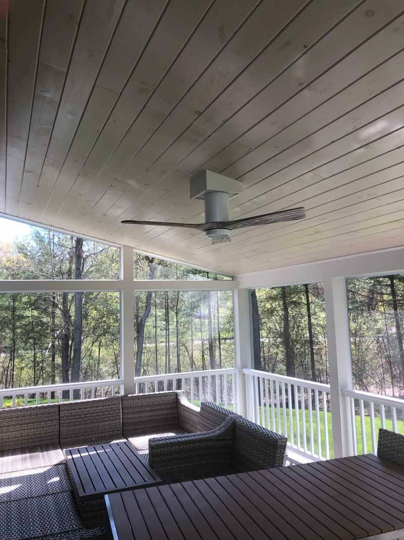 Covered patio with a ceiling fan, wicker furniture, and a view of trees through screened windows.