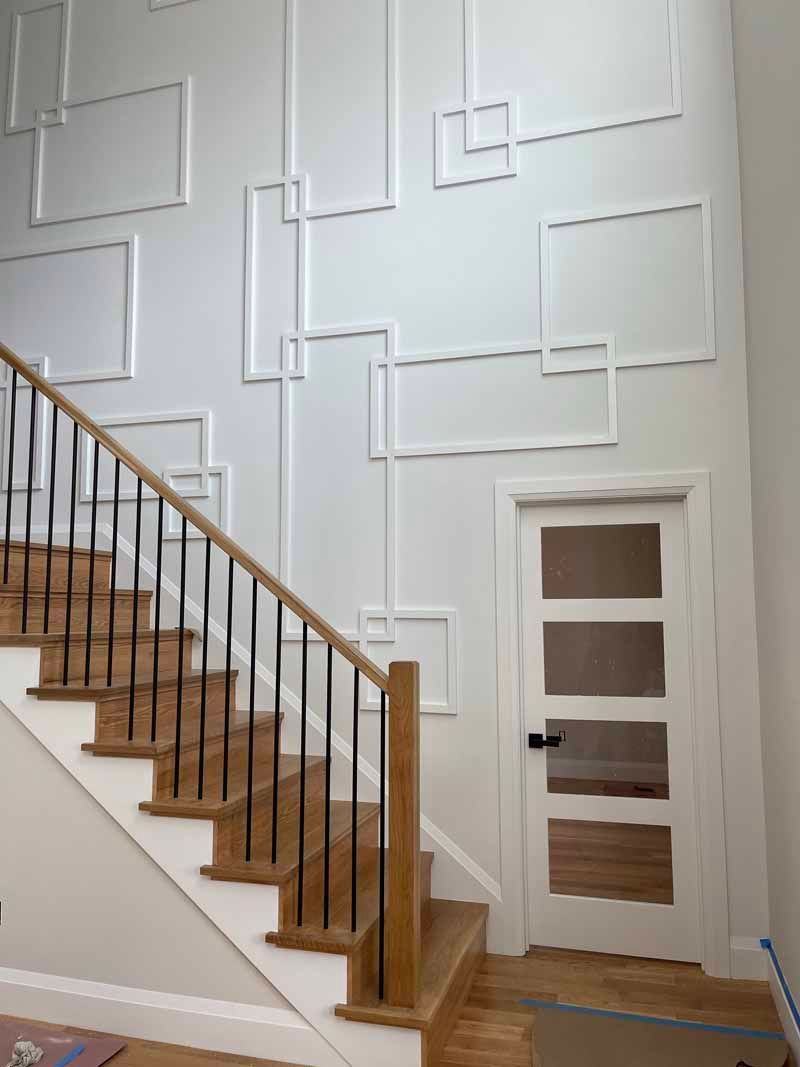 Staircase with wooden steps and black railings, white wall with geometric molding, and a white door with glass panels.