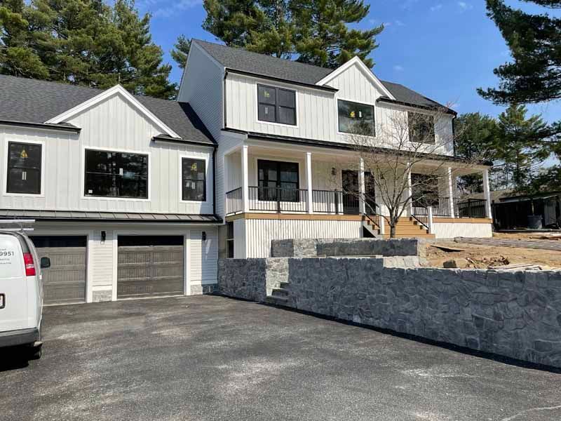 White farmhouse with black trim, gray stone retaining wall, two-car garage, and asphalt driveway.