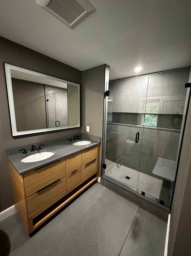 Modern bathroom with gray tile, wood vanity, glass shower, and rectangular mirror.