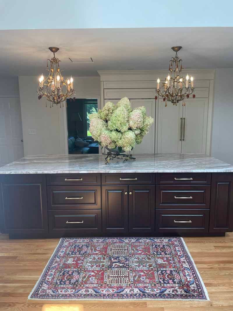 Kitchen island with dark cabinetry, marble countertop, and chandeliers. Hydrangeas sit on the island, and a rug on the hardwood floor.