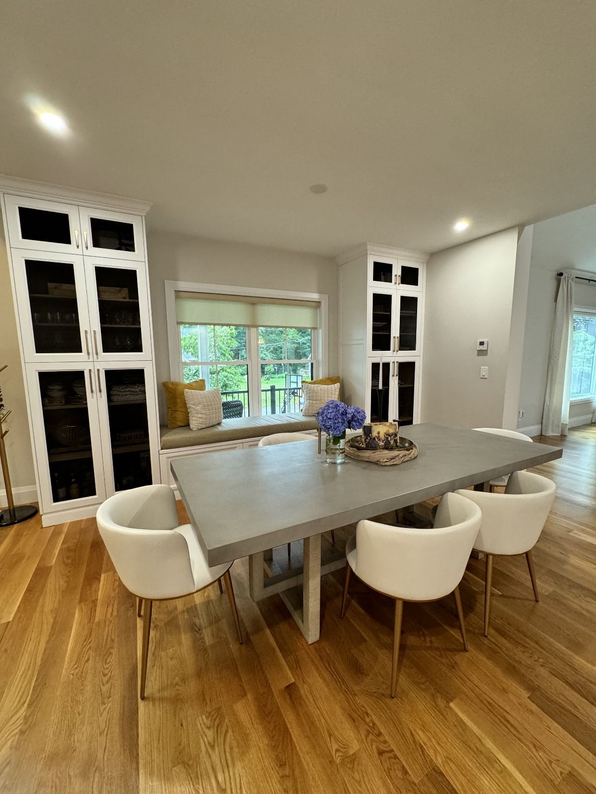 Dining room with concrete table, white chairs, built-in cabinets, and a window seat with pillows.