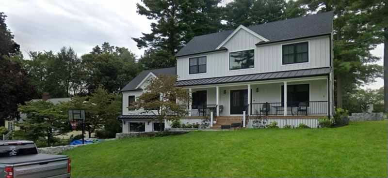 White farmhouse with black trim and a porch, set on a green lawn, surrounded by trees.