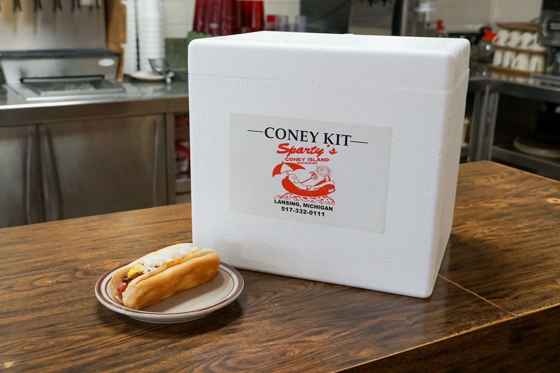 A hot dog on a plate next to a styrofoam box that says coney kit