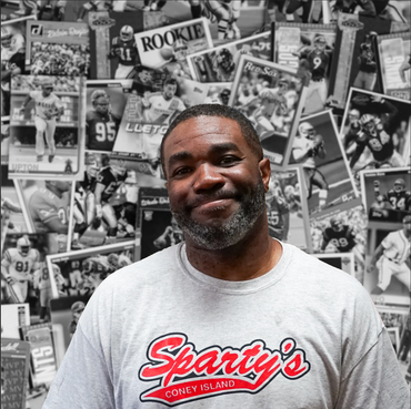 A man wearing a sparty 's shirt stands in front of a wall of football cards