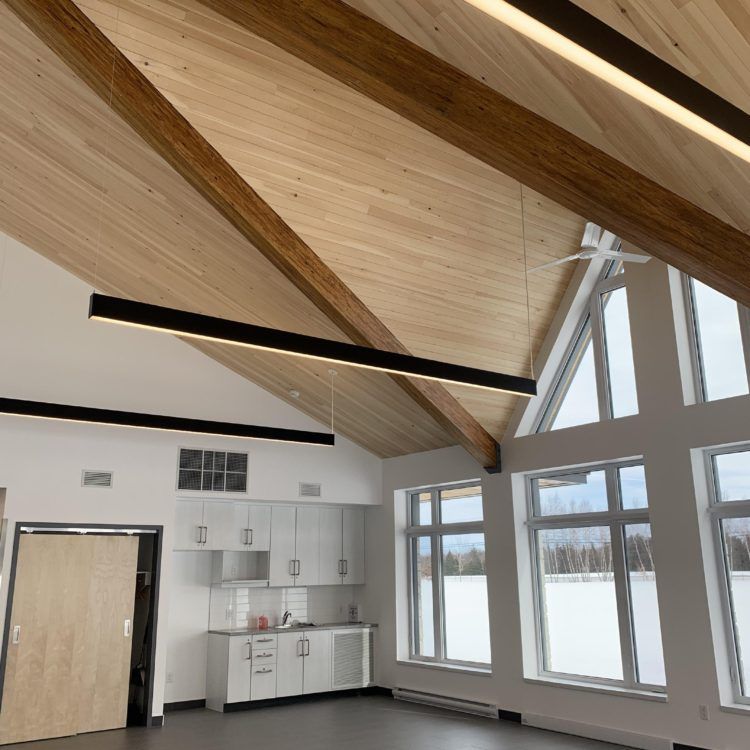 Vue intérieure d'un bâtiment avec un plafond incliné lambrissé, des poutres apparentes et de grandes fenêtres.