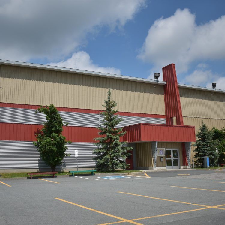 Vue extérieure d'une arène avec une façade rouge et beige, une entrée, des arbres et un parking.