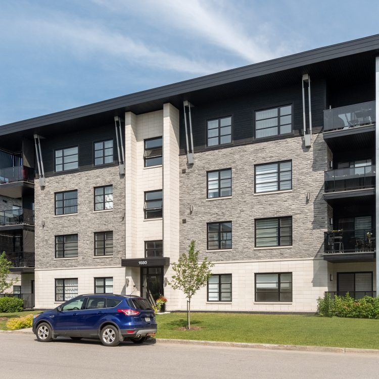 Un SUV bleu est garé devant un immeuble d'appartements moderne de trois étages, avec des éléments en pierre et des touches de noir.