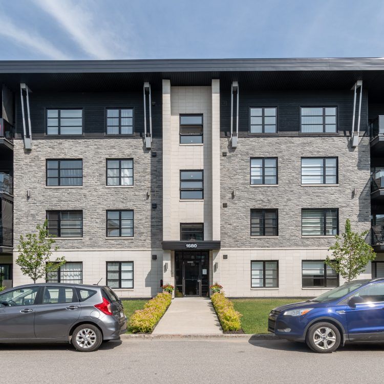 Extérieur d'un immeuble d'appartements, façade en pierre grise, voiture bleue et voiture grise garées devant.