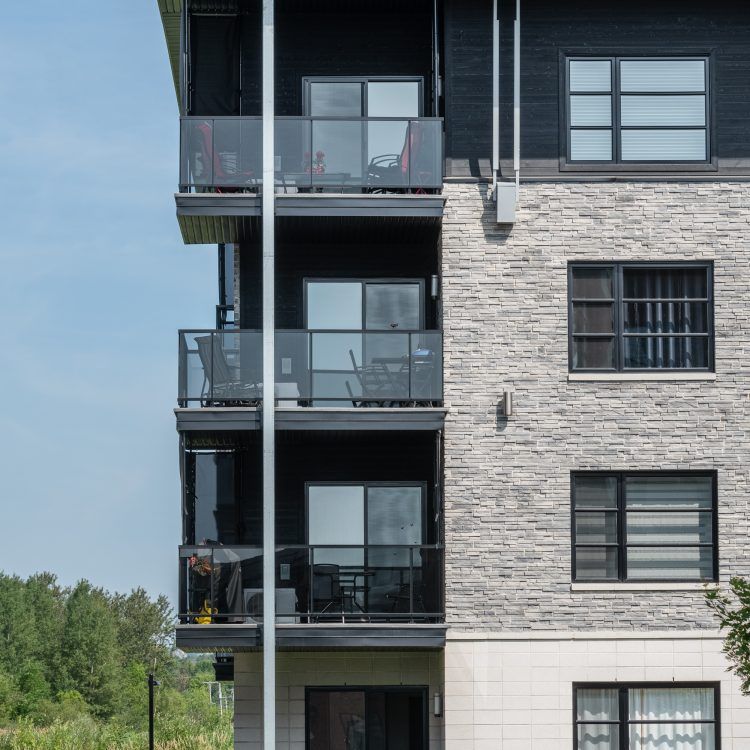 Immeuble à plusieurs étages avec balcons ; façade en pierre grise, bardage en bois foncé, ciel bleu.