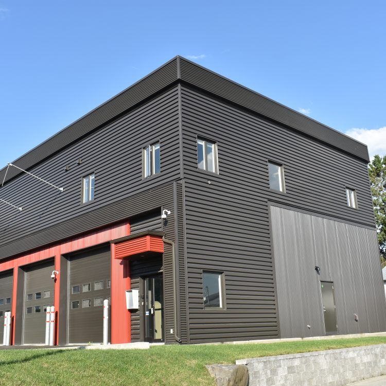 Bâtiment noir et rouge avec portes de garage, petites fenêtres et pelouse sous un ciel bleu.
