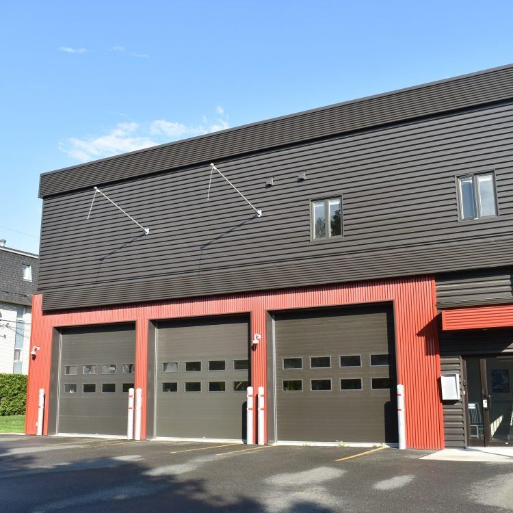 Caserne de pompiers avec trois portes de garage. Façade noire au-dessus de portes rouges et grises, sous un ciel bleu.