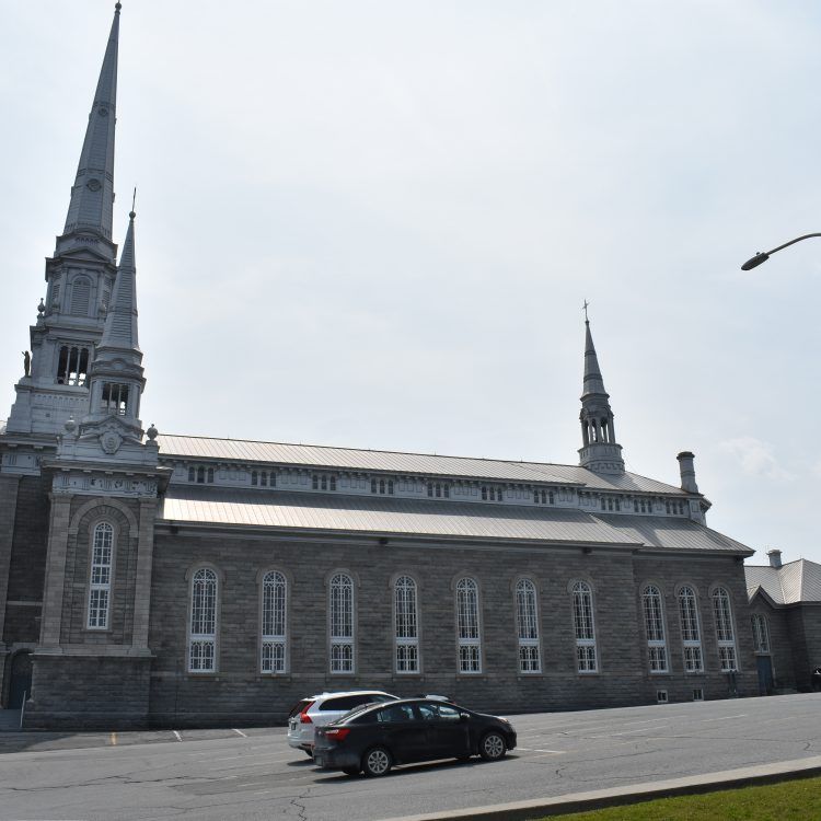 Église en pierre avec de hauts clochers, une façade en pierre grise, des fenêtres cintrées et deux voitures garées devant.