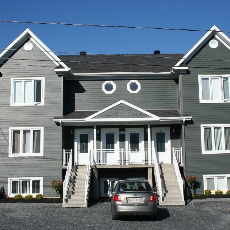 Immeuble d'appartements gris de deux étages avec deux portes d'entrée et une voiture garée devant.