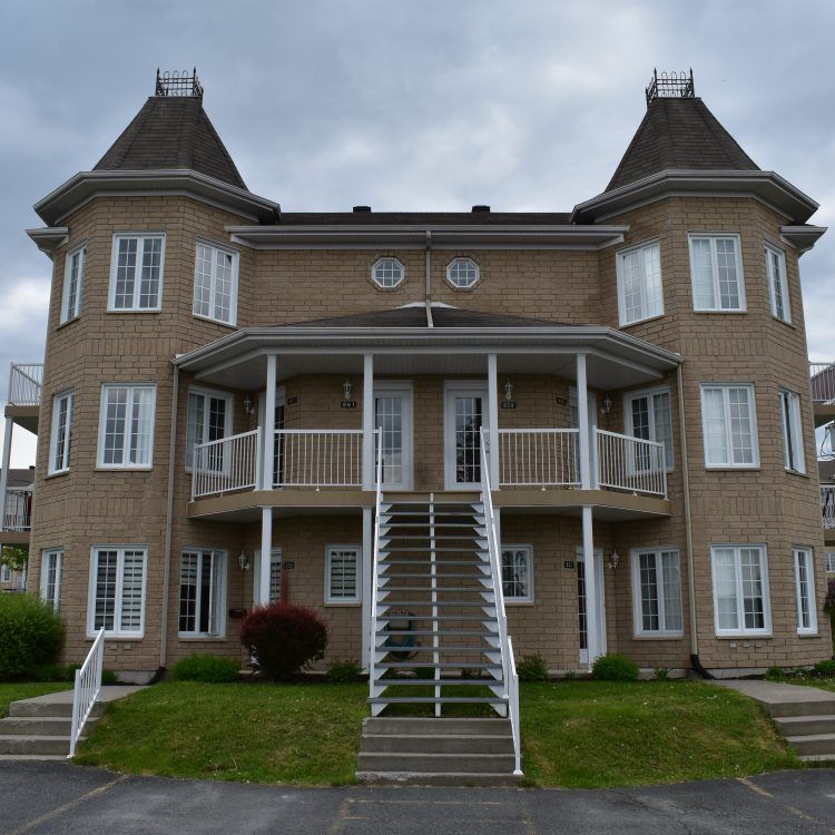 Bâtiment en briques de deux étages avec deux tourelles jumelles. Balcons, fenêtres et escaliers blancs. Ciel couvert.