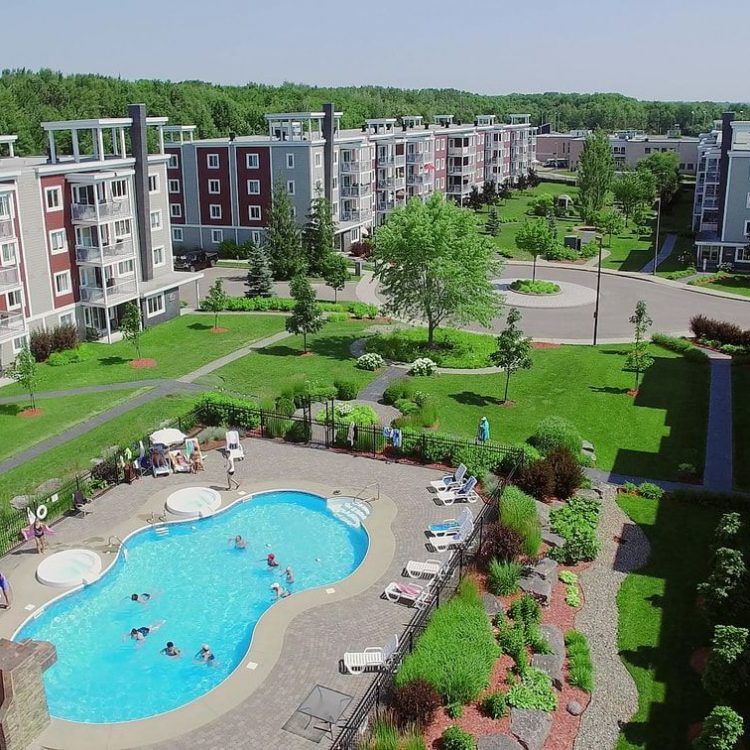 Vue aérienne d'un complexe d'appartements avec piscine, pelouses verdoyantes et arbres par une journée ensoleillée.