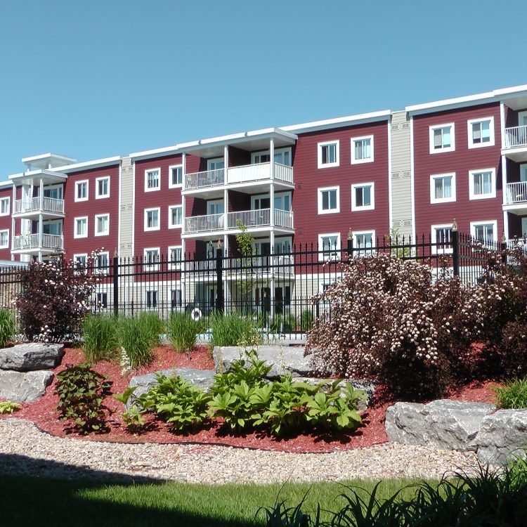 Immeuble d'appartements avec bardage bordeaux, boiseries blanches et balcons, derrière une clôture noire et des aménagements paysagers.