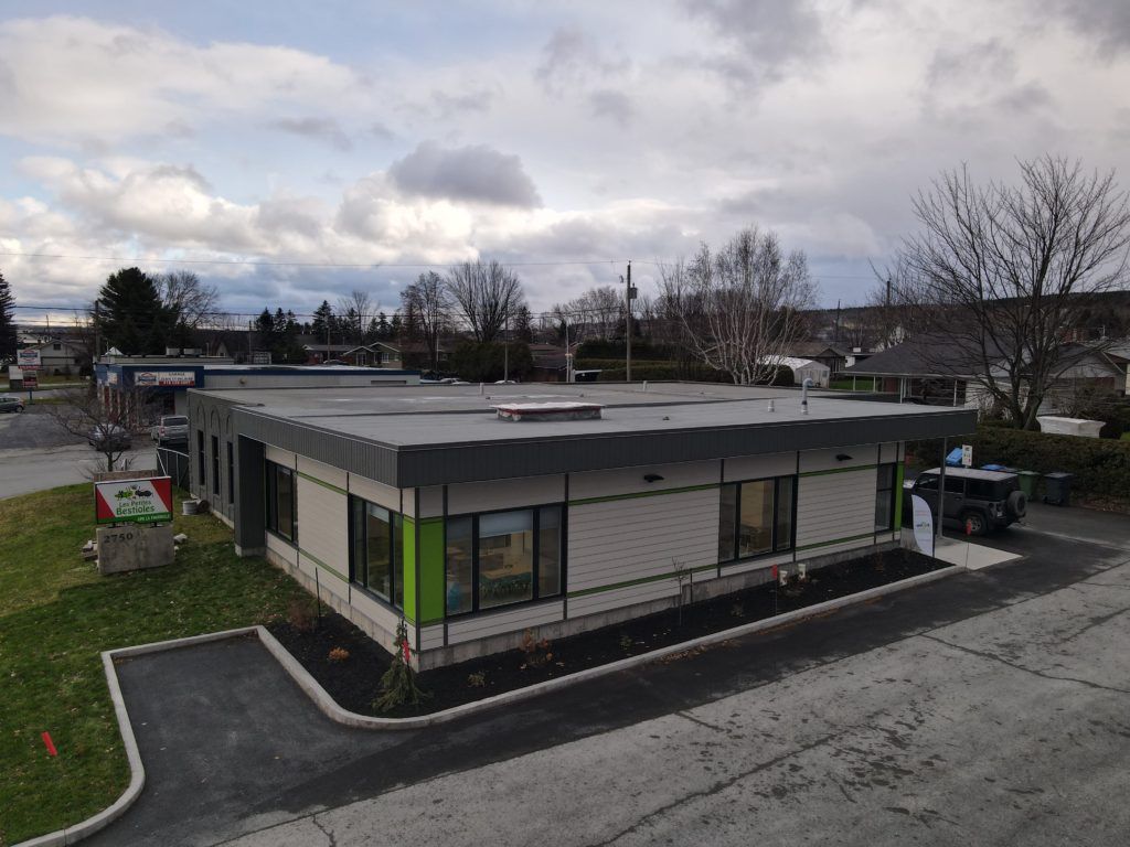 Bâtiment moderne de plain-pied avec toit plat. Fenêtres, touches de gris et de vert, et parking goudronné. Ciel nuageux.