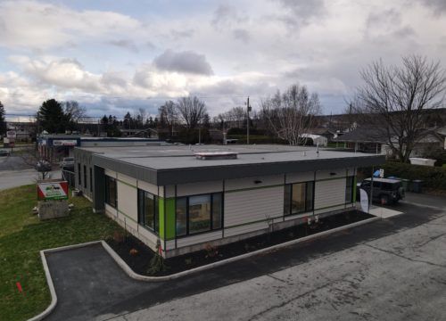 Bâtiment moderne de plain-pied avec toit plat ; bardage beige, touches de vert et de gris, fenêtres. Ciel nuageux.