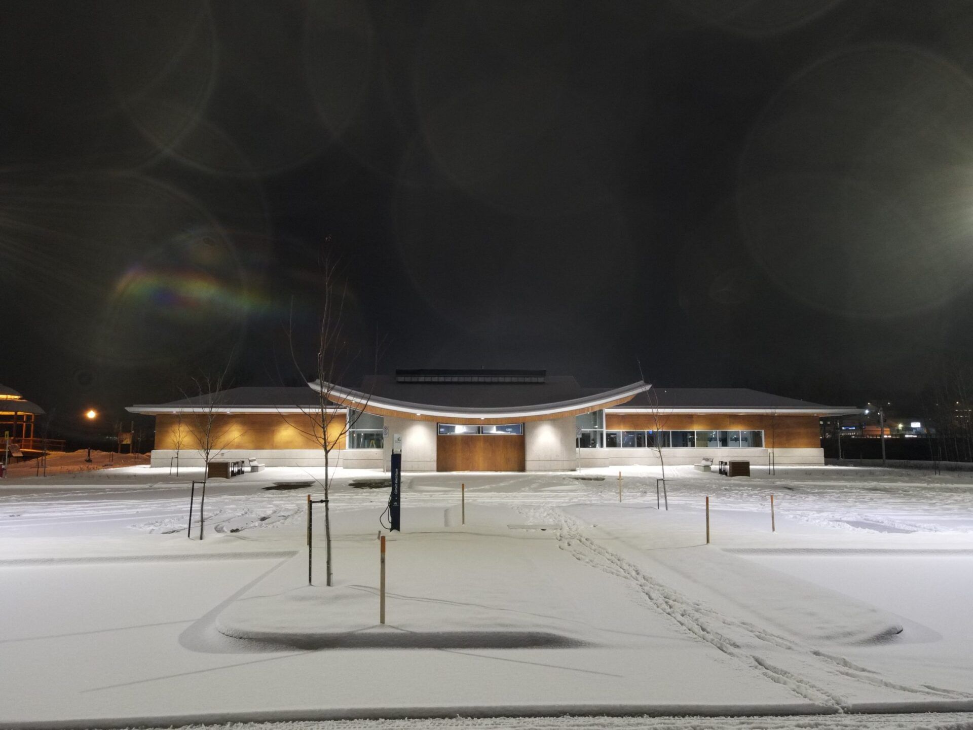 Bâtiment enneigé la nuit ; toit incurvé, façade en bois, intérieur éclairé. Empreintes de pas dans la neige menant à l’entrée.