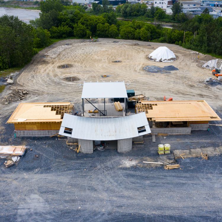 Chantier de construction avec une structure partiellement édifiée, entouré de gravier et de terrain nu, avec des arbres et des bâtiments en arrière-plan.