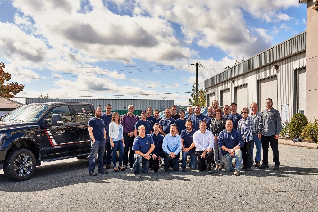 Un groupe de personnes pose en extérieur devant un bâtiment et un camion noir, sous un ciel nuageux.