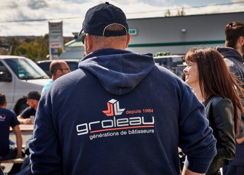 Un homme en sweat-shirt bleu à capuche avec le logo « Groleau », dos à la caméra, discute avec une femme souriante à l'extérieur.