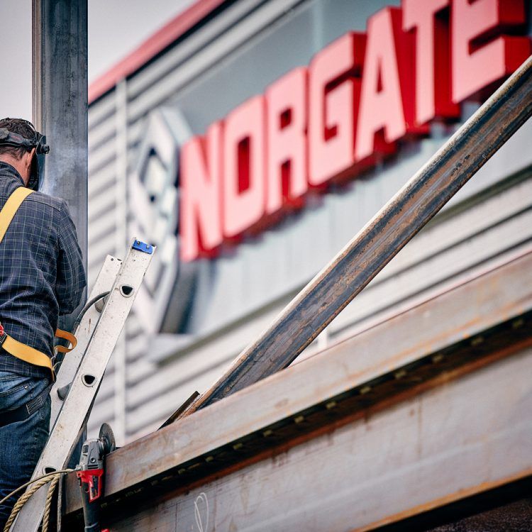 Un soudeur travaille sur une structure métallique à l'extérieur d'un bâtiment portant l'enseigne « NORGATE » en rouge.