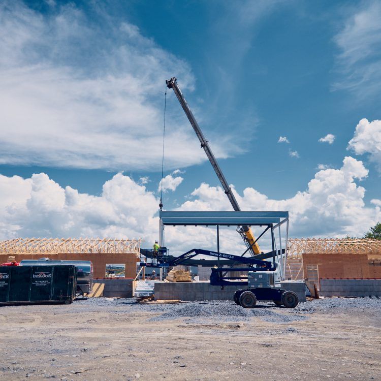 Chantier de construction avec une grue soulevant des poutres métalliques pour la charpente d'un bâtiment. Ciel bleu, nuages ​​et ouvriers sont visibles.