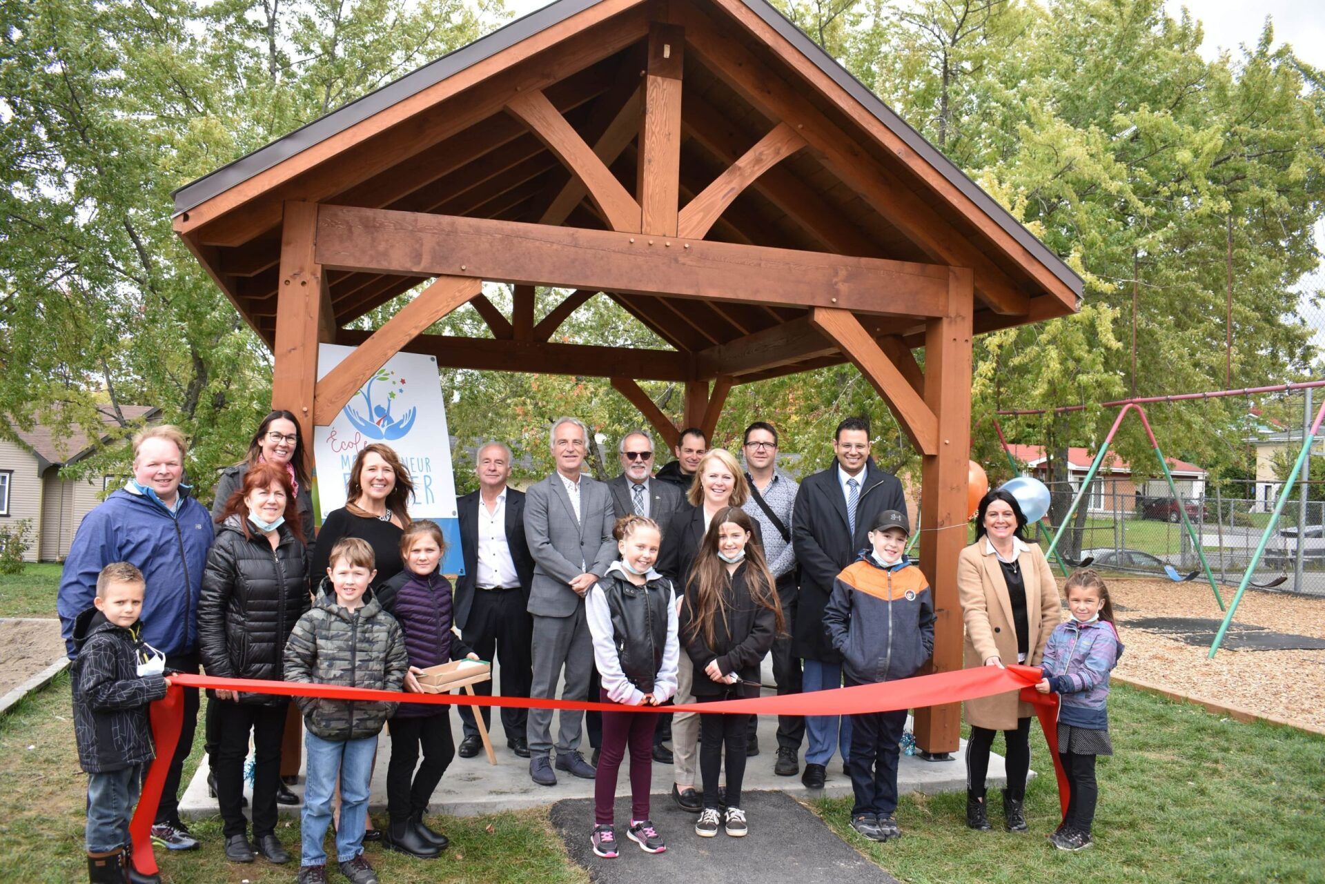 Un groupe coupe un ruban rouge devant un kiosque en bois dans un parc ; enfants et adultes sourient.