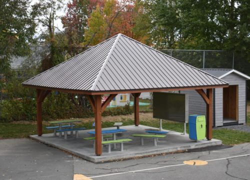 Un espace extérieur couvert avec des tables de pique-nique, un grand tableau noir et un toit en métal.