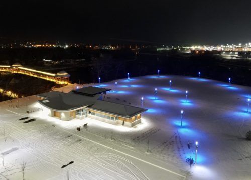 Vue aérienne nocturne d'un paysage enneigé avec un bâtiment éclairé et des poteaux illuminés en bleu ; des arbres au loin.
