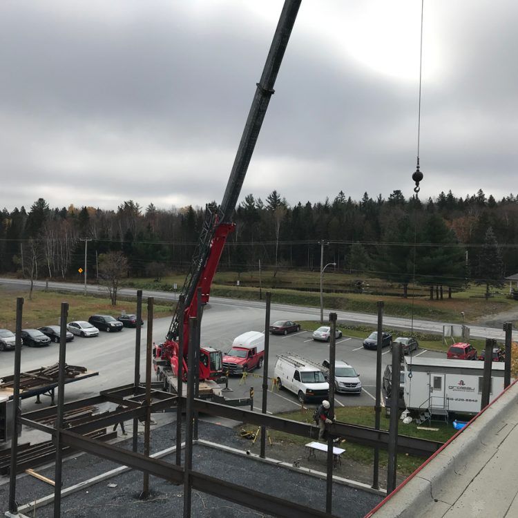 Chantier de construction avec grue soulevant des poutres d'acier ; un ouvrier est présent. Ciel couvert.
