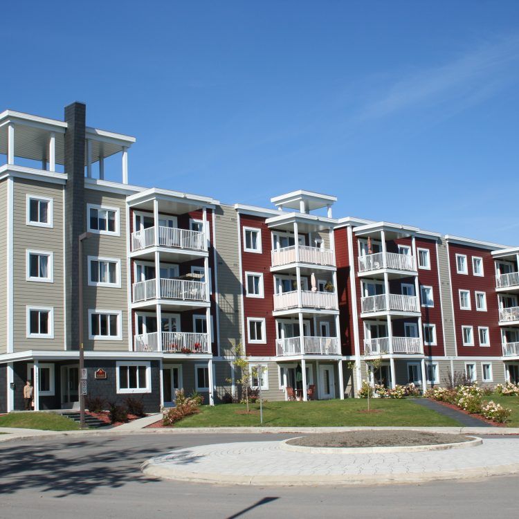 Immeuble d'appartements à plusieurs étages, façade grise et rouge, balcons blancs, allée circulaire, ciel bleu.