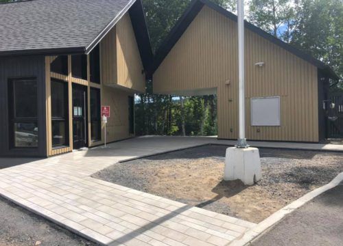 Un bâtiment bas avec une rampe et un mât de drapeau. Bardage en bois, pavés gris et forêt en arrière-plan.