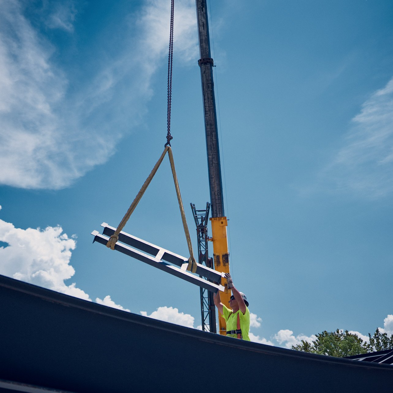 Une grue soulève une poutre métallique pour la poser sur un toit ; un ouvrier en gilet jaune observe la scène. Ciel bleu nuageux en arrière-plan.