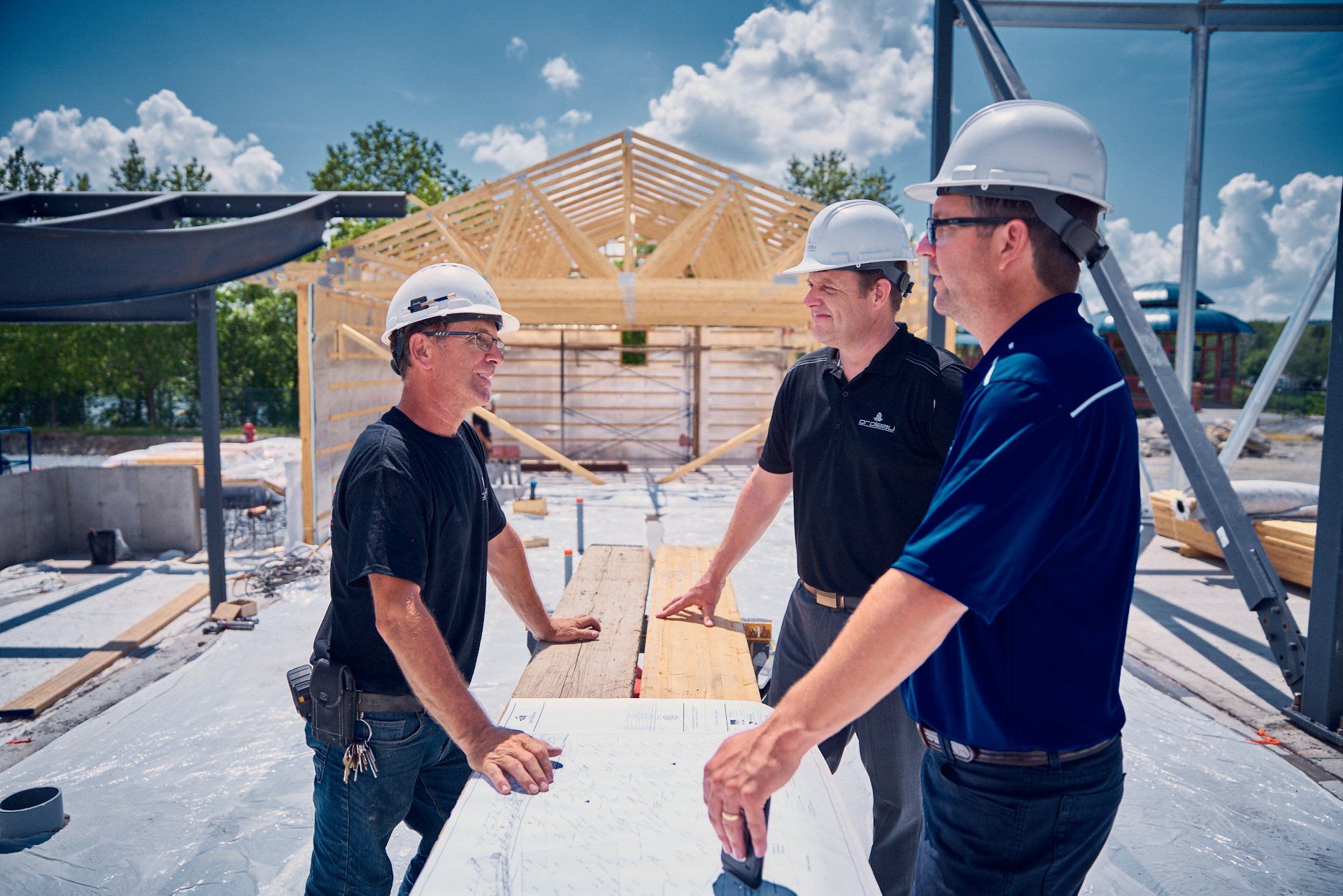 Trois ouvriers du bâtiment, casqués, discutent des plans sur un chantier. Ciel bleu clair visible.