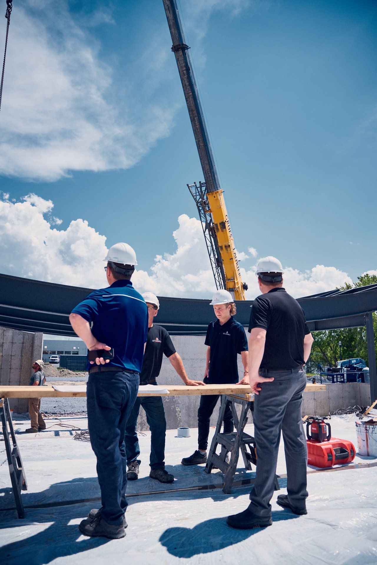 Des ouvriers du bâtiment, casqués, examinent des plans près d'une grue sous un ciel bleu.