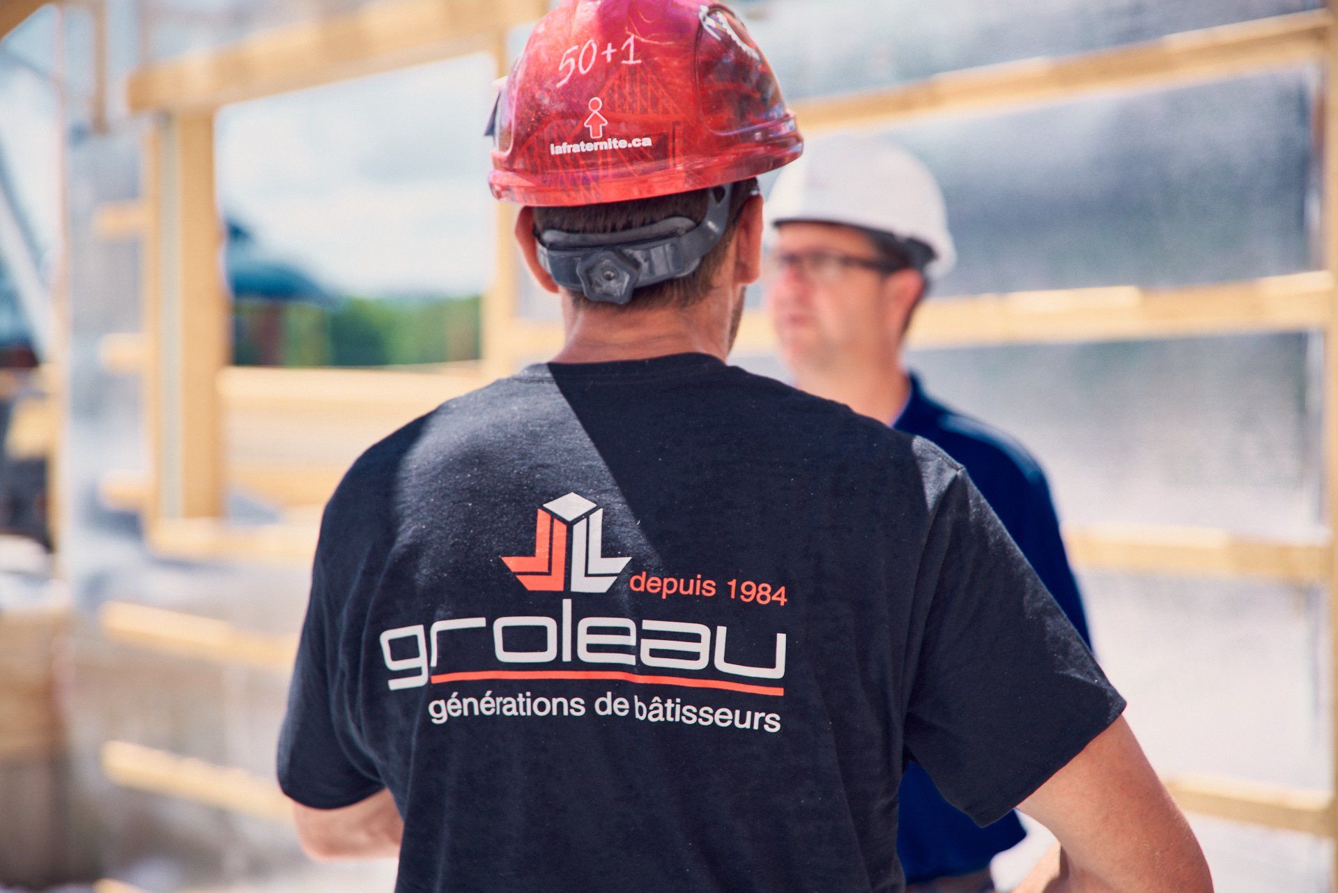 Deux ouvriers du bâtiment, dont l'un porte un casque rouge, discutent sur un chantier. Logo « Groleau » sur sa chemise.