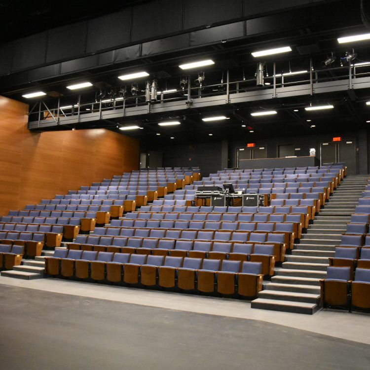 Théâtre vide avec des rangées de sièges bleus en gradins et un éclairage orienté vers la scène.