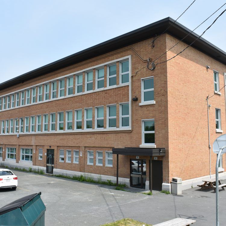 Bâtiment scolaire en briques de deux étages avec de nombreuses fenêtres et un petit auvent à l'entrée ; vue extérieure.