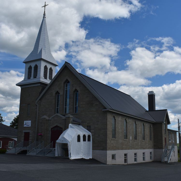 Église en pierre au clocher élancé, sous un ciel bleu parsemé de nuages. Un voile blanc se trouve près de l'entrée.