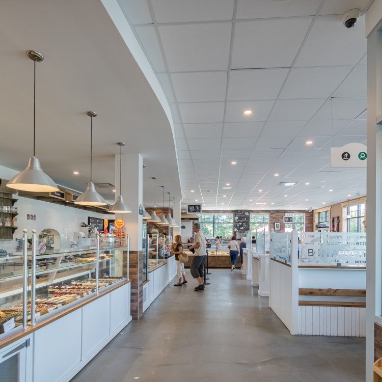 Intérieur d'une boulangerie avec des clients. Vitrines de pâtisseries, murs blancs et suspensions lumineuses.