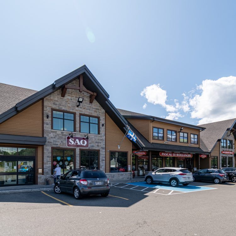 Vue extérieure d'un centre commercial avec divers magasins, des voitures garées et un ciel bleu.