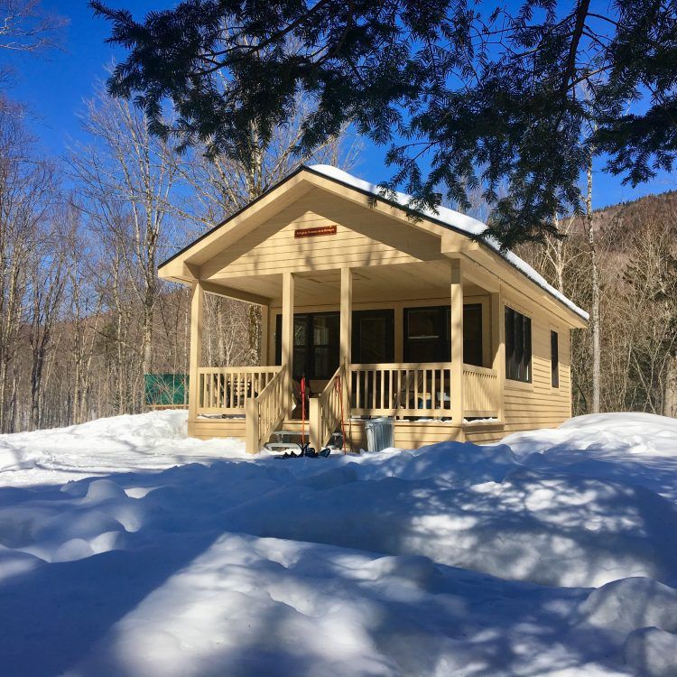 Petite cabane enneigée avec une véranda, entourée d'arbres sous un ciel bleu clair.