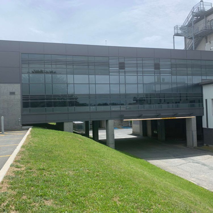 Bâtiment moderne avec une passerelle vitrée surplombant une pente herbeuse et une zone pavée.