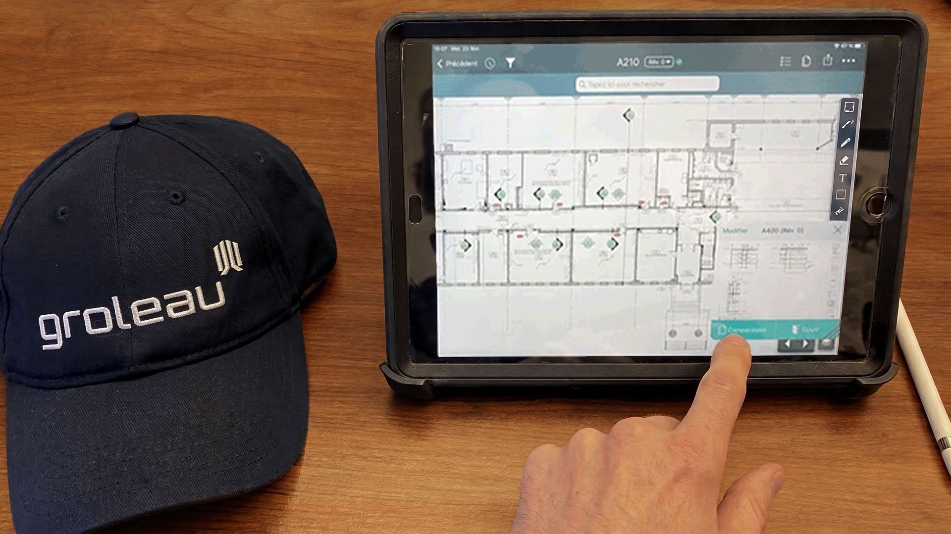 La main d'une personne touche une tablette sur laquelle est affiché un plan d'étage ; une casquette de baseball est posée à proximité sur une table.